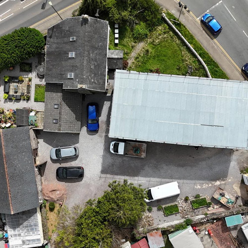 aerial view of a residential property with multiple vehicles parked in a gravel lot surrounded by greenery and a metal roof highlighting the landscape and structure design