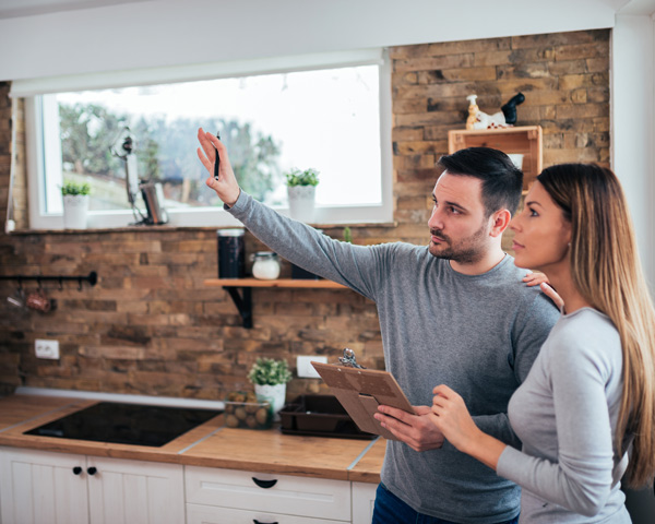 man in gray shirt and woman discussing kitchen renovation while holding clipboard in modern kitchen with wooden accents and plants