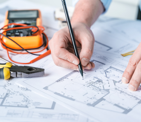 hand holding a pencil drawing electrical plans on blueprints with tools and a multimeter in the background showcasing electrical system designs for construction plans 4 innovative ideas for projects